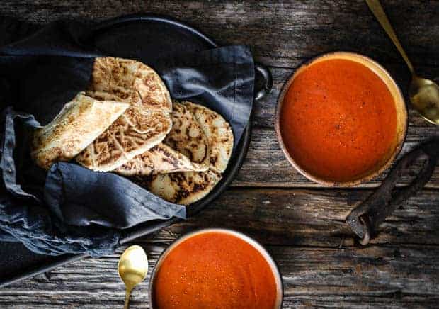 a basket with a gray linen napkin is full of golden paleo naan. There are two bowls of tomato soup next to the basket of naan bread.