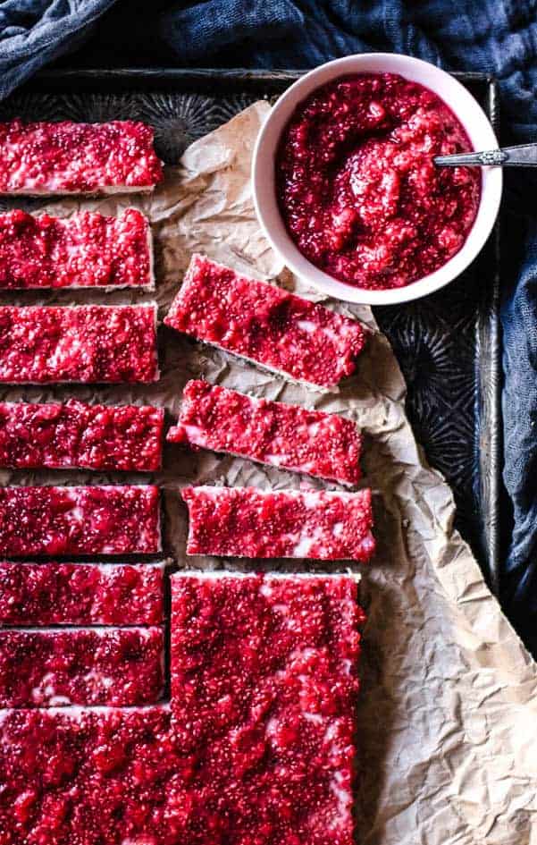 12 coconut bars layered with raw raspberry jam sit on a large piece of brow parchment paper. there is a sheet pan underneath the parchment and a bowl of raw raspberry jam next to the bars.