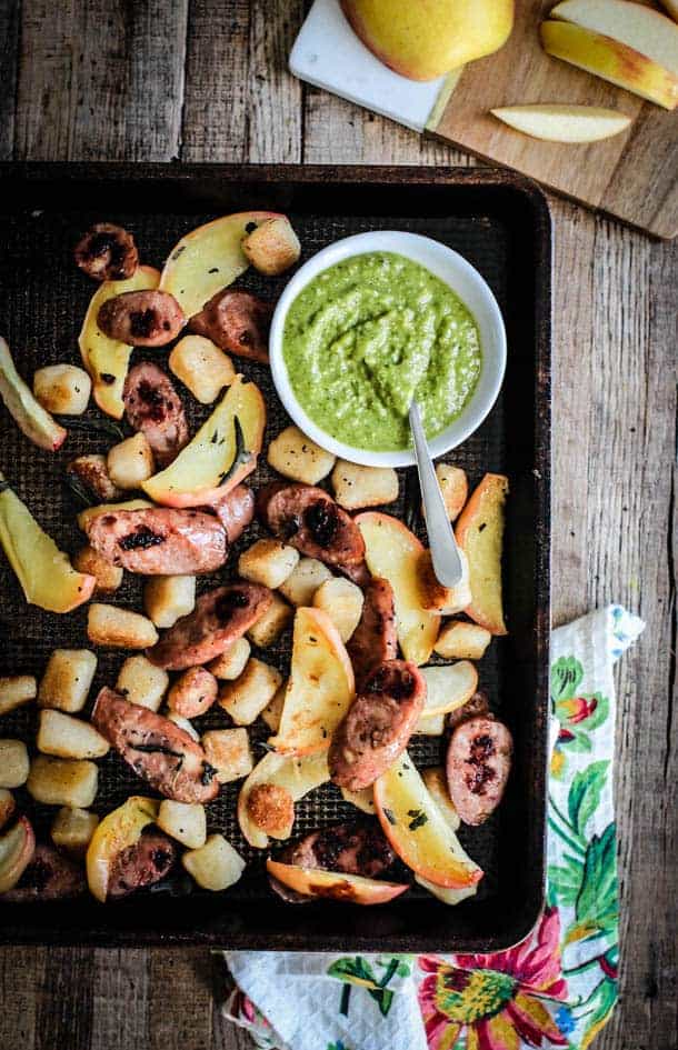 A sheet pan full of roasted cauliflower gnocchi, apples, sage leavs, and sausage slices. A bowl of brigt green pesto sits in the sheetpan as well.