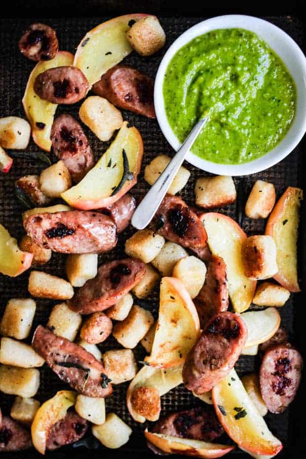 A close up of a sheet pan full of rpasted cauliflower gnocchi, apple slices, sausage slices, and sage leaves. there is a small bowl of pesto on the sheet pan for serving.