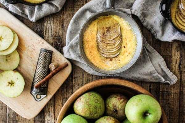 a bowl of green apples and a curling board sit on a table next to a skillet with baked apple custard inside. 