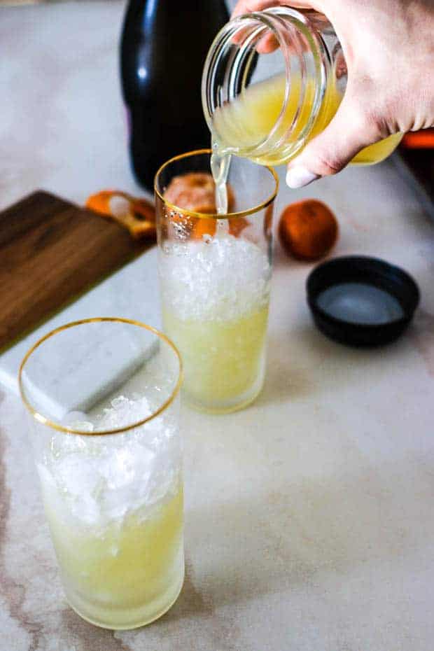 2 cocktail glasses are filled with ice and a woman is pouring the mandarin basil and ginger simple syrup into them.