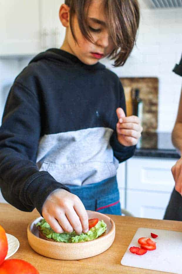 A boy decorating a slice of fruity avocado toast with sliced berries 