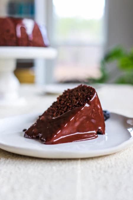 A slice of Chocolate Covered Prune Fudge Cake on a plate in front of the rest of the cake on a cake stand