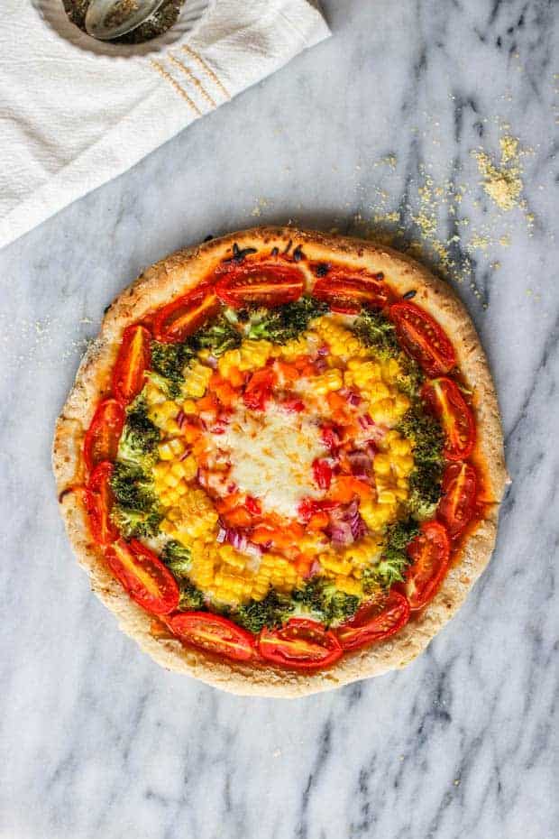 An entire pizza is on a marble surface. The pizza is a vegetable pizza it has tomatoes, broccoli, corn, orange bell peppers, and red onions have been arranged in circles around the dough to create a circular rainbow. 