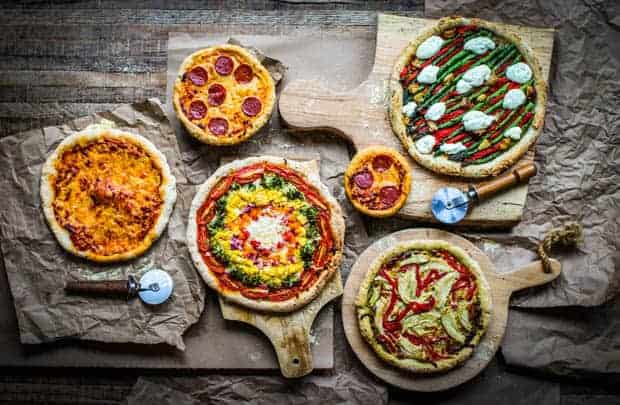6 pizza arranged on a table, there is a rainbow veggie pizza, cheee pizza, pepperoni pizza, asparagus goat cheese pizza, and chicken pesto pizza