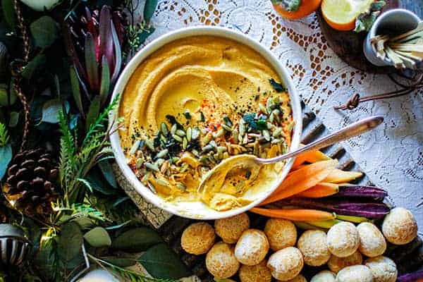 A bowl of Roasted Butternut Hummus with Crispy Fried Sage sprinkled over the top served with brazilian cheese bread and rainbow carrots