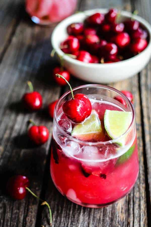 A bright red Fresh Cherry Limeade Cocktail garnished with lime quarters and fresh cherries on a table next to a bowl of fresh cherries