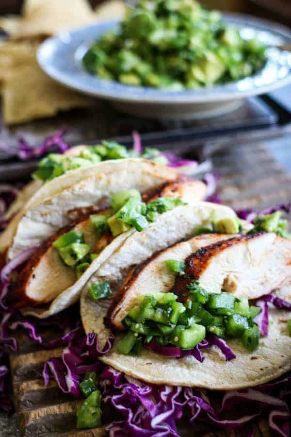 3 Easy Grilled Enchilada Chicken tacos on a platter next to a bowl of green salsa. the tacos gave purple cabbage, sliced grilled chicken, and green pico de gallo