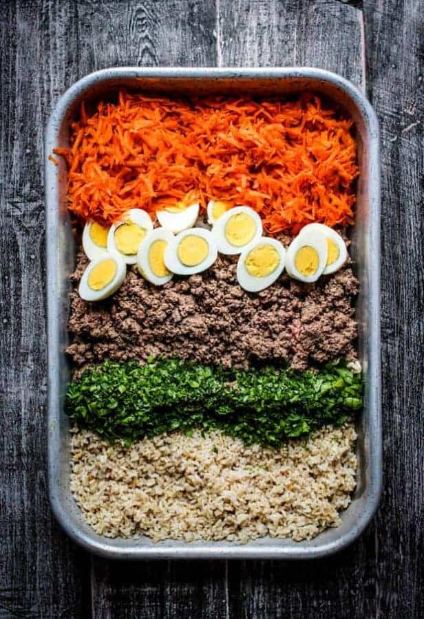 All of the ingredients for homemade dog food in a baking dish before being mixed together. Ground beef, brown rice, shredded carrot, chopped hard boiled eggs, and minced parsley