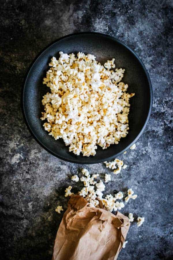 A brown bag of popcorn spilling out onto a table next to a big bowl of popcorn.