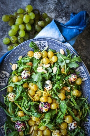 A platter filled with Roasted Grape & Arugula Salad - roasted green grapes, arugula, goat cheese balls rolled in edible flowers, and slivered almonds