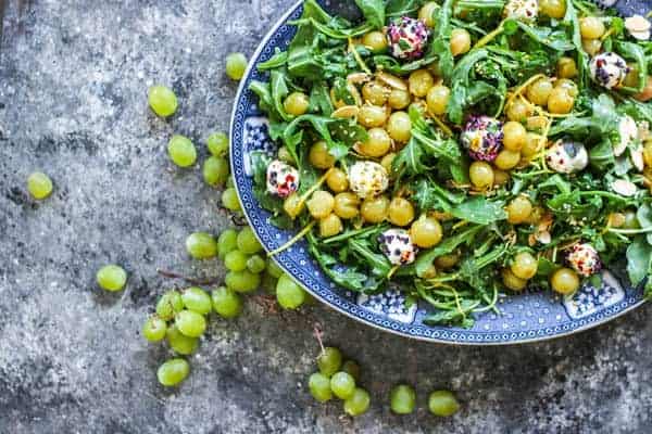A platter of Roasted Grape & Arugula Salad with green grapes on the table next to it. 