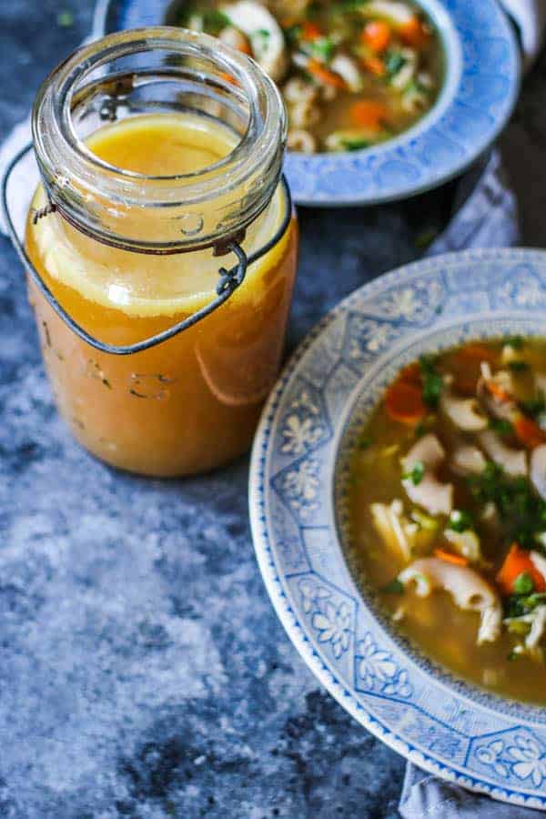 A jar of The Easiest DIY Rotisserie Chicken Broth on a table next to a bowl of chicken noodle soup