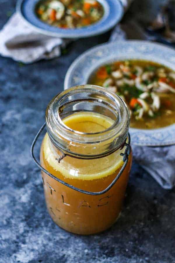 A jar of The Easiest DIY Rotisserie Chicken Broth is sitting on a table with bowl of homemade chicken noodle soup.