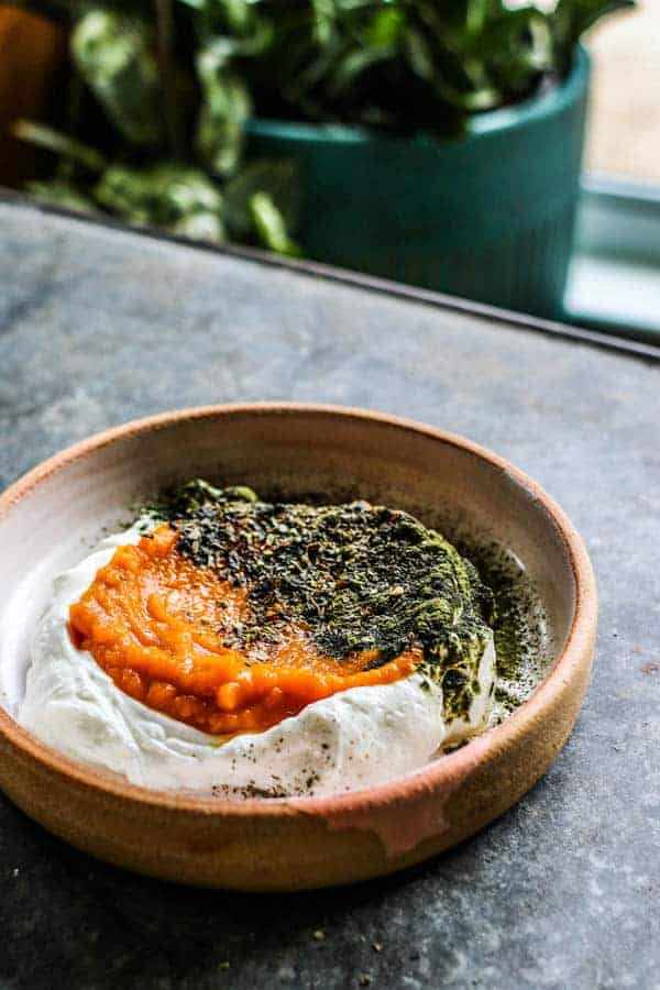 A Pumpkin and Yogurt Bowl for Dogs sits on a table. It is layered with greek yogurt, pumpkin puree, and a sprinkling of green vitamin powder and dried herbs