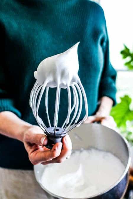 A woman in a green sweater holding a kitchen aid stand mixer bowl and whisk attachment with aquafaba on the end. She is showing that aquafaba whips up like egg whites. 