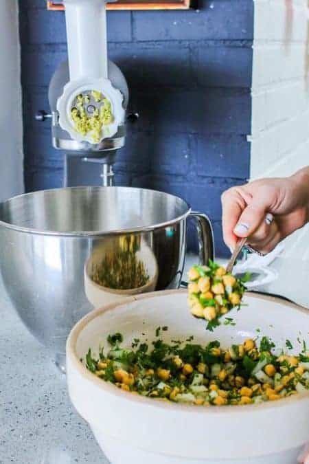 A woman spooning a mixture of soaked chickpeas and herbs into a meat grinder attached to a Kitchen aid stand mixer