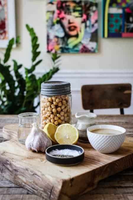 A wooden cutting board that has a lemon half, garlic bulb, a glass jar of chickpeas, and bowl of tahini on top.