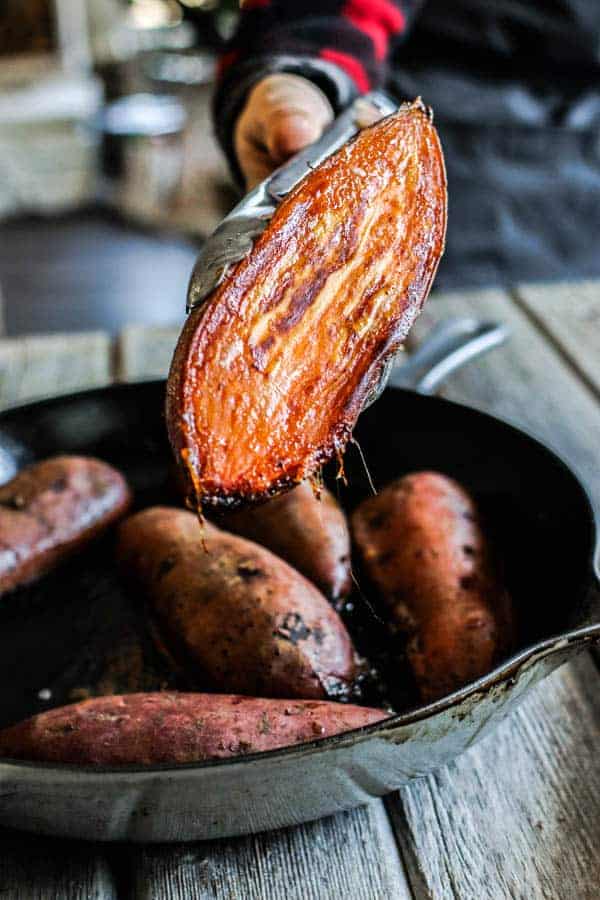 A woman holding a halved, roasted sweet potato with tongs towards the camera. The bottom of the sweet potato is caramelized from roasting in the butter