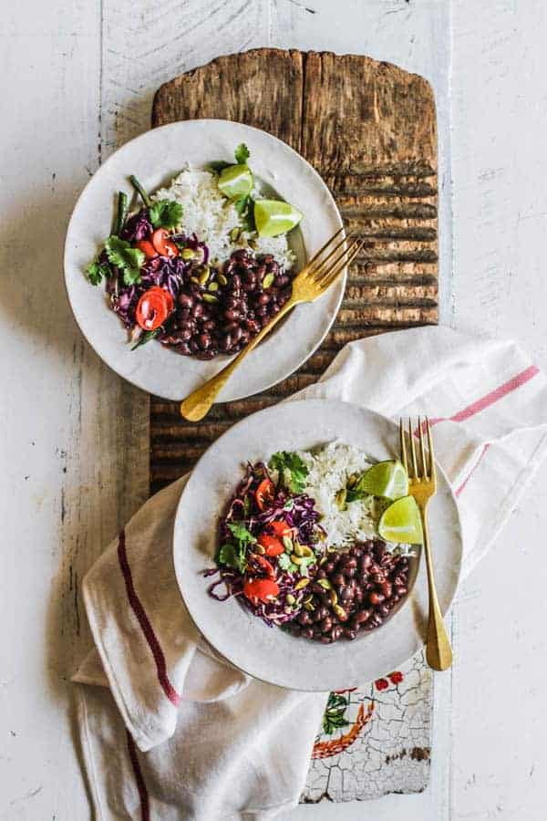 Two burrito bowls on a table with white rice, black beans spicy colorful slaw, cilantro, and lime wedges