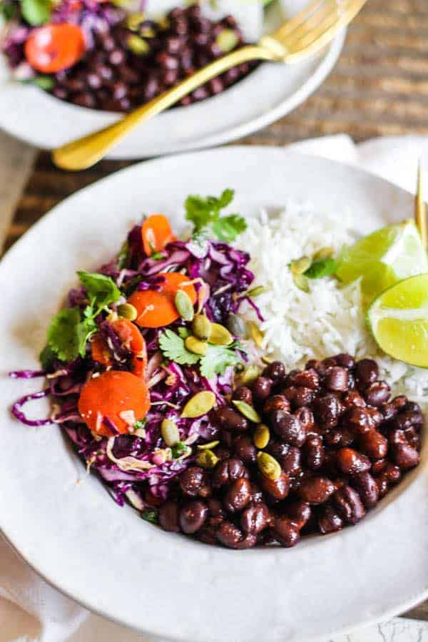 A bowl of black beans with rice, spicy colorful slaw, cilantro and lime wedges.