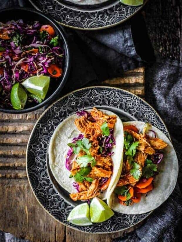 Two Shredded Instant Pot Mexican Chicken recipe tacos are on a plate with lime wedges next to a bowl of colorful spicy slaw