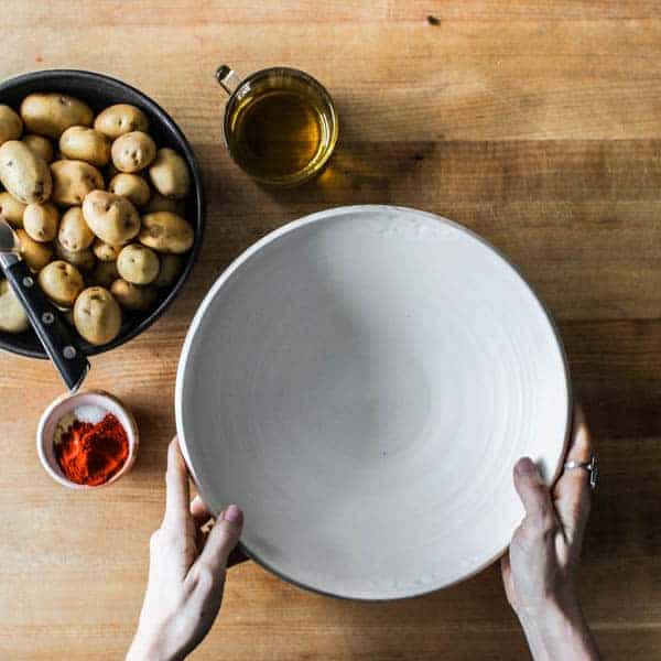 An empty mixing bowl, a smaller bowl full of baby yellow potatoes. a tiny bowl of red paprika, and a measuring cup of oil are on a chopping block