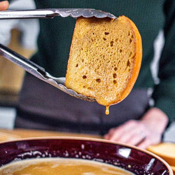 A slice of brioche that has been dunked in the French toast custard and the excess is running off while it is being held in mid air by tongs.  