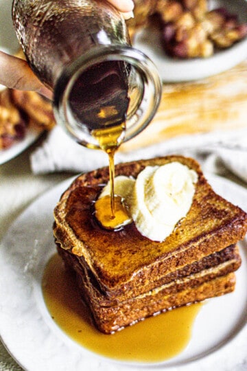 Slices of french toast stacked on a white plate with sliced bananas on top. maple syrup is being poured over the top of the French toast.