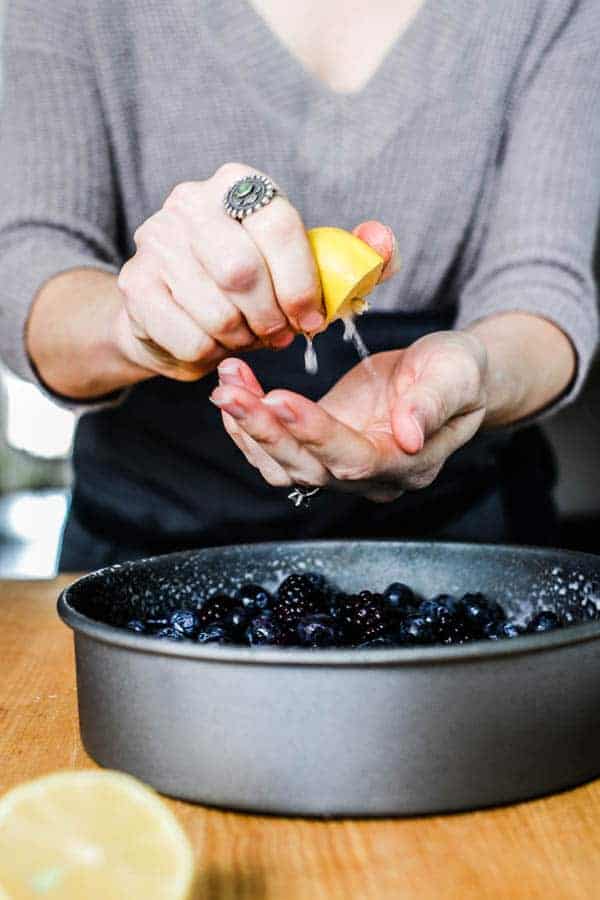 A woman squeezing a lemon over a baking pan full of berries. She is making a black and blueberry cobbler.
