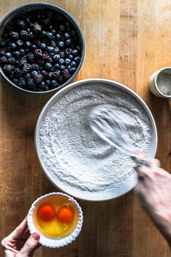 A bowl of flour and sugar being whisked . Small bowls of eggs and berries sit around the bowl of flour. The ingredients will bake up to make a black and blueberry cobbler.