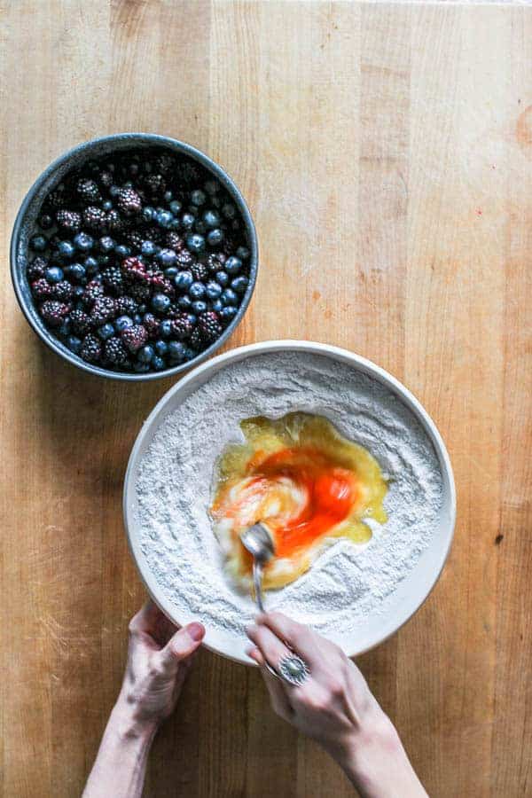 Eggs being whisked into dry ingredients next to a bowl of blueberries. This will become the batter for black and blueberry cobbler.