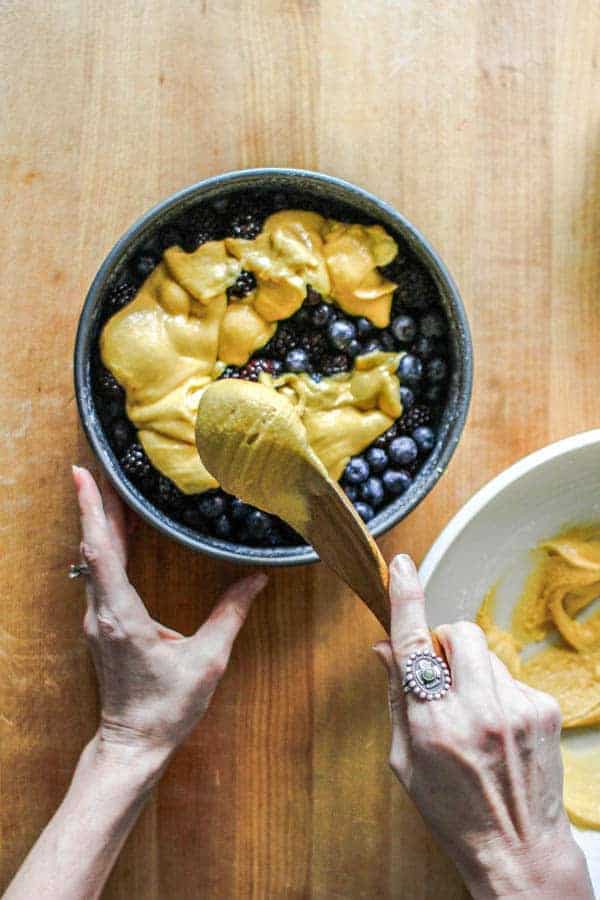 The batter for the crust of a black and blueberry cobbler being spread over berries in a baking pan. 