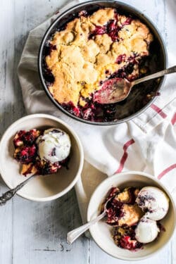 A pan of freshly baked black and blueberry cobbler next to bowls that have been served with cobbler and vanilla ice cream.