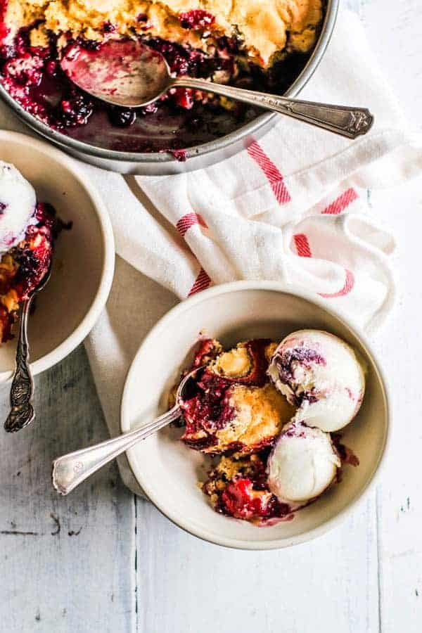 2 bowls of black & blueberry cobbler with ice cream next to a pan of cobbler 