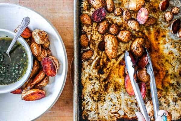 A sheet pan sits on a counter with oven roasted potatoes on it and a pair of tongs. There is a bowl next to the sheet pan that has a few scoops of the oven roasted potatoes in it and a smaller bowl that has a green dipping sauce