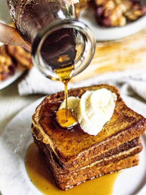 Slices of french toast stacked on a white plate with sliced bananas on top. maple syrup is being poured over the top of the French toast.