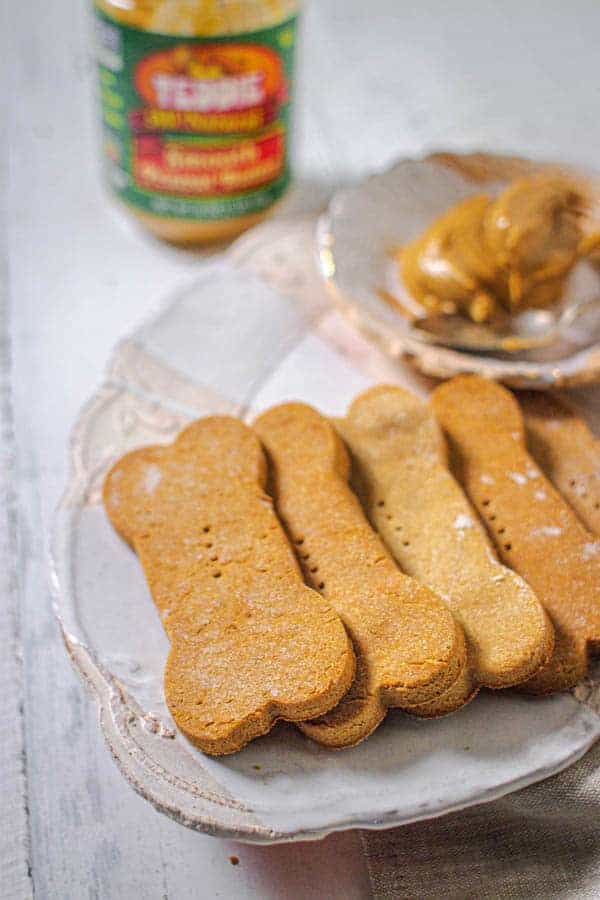 Homemade peanut butter dog treats shaped like bones on a plate next to a bowl of peanut butter.