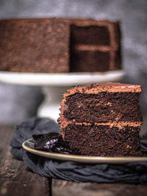 A 2-layer chocolate cake with chocolate icing, decorated with chocolate sprinkles is in the background while a slice of the cake is plated in front with a few Californian prunes on the plate.