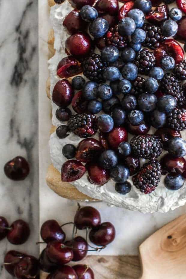 A top down view of a red, white, and blue dessert cake topped with mascarpone  whipped cream, pitted cherries and fresh blueberries and blackberries. 
