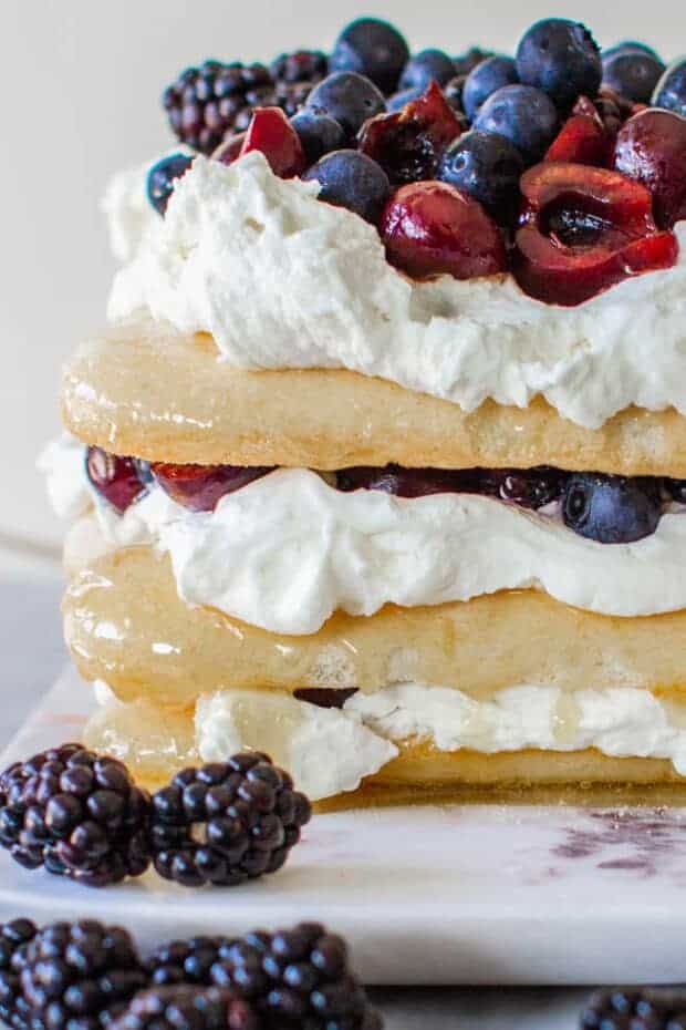 This red, white, and blue dessert cake from the side view. Stacked citrus-caramel soaked Lady Fingers, layered with with mascarpone whipped cream and fresh cherries, blackberries and blueberries