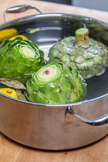 Three prepared artichokes swimming in a large pot filled with water, lemon halves, & bay leaves.