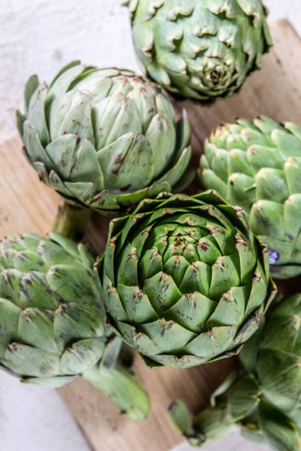 Whole artichokes stacked on a wooden cutting board