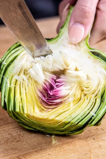 Half of a globe artichoke wuith teh fuizzy choke being removed with a pairing knife.
