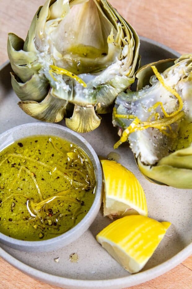 One artichoke cut in half on a grey plate. They are topped with salt,pepper & lemon peel curls. In a dish to the side there is an olive oil dipping sauce with salt,pepper, & lemon peel curls aswell. Two lemon slices for garnish on the side of the sauce bowl.