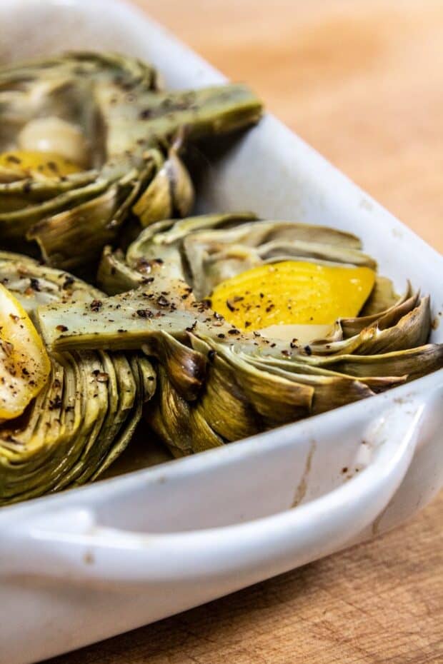 Artichoke halves placed in a a white baking dish with the middle facing up. The artichokes have lemon slices & a clove of garlic in the middle topped off with salt & pepper