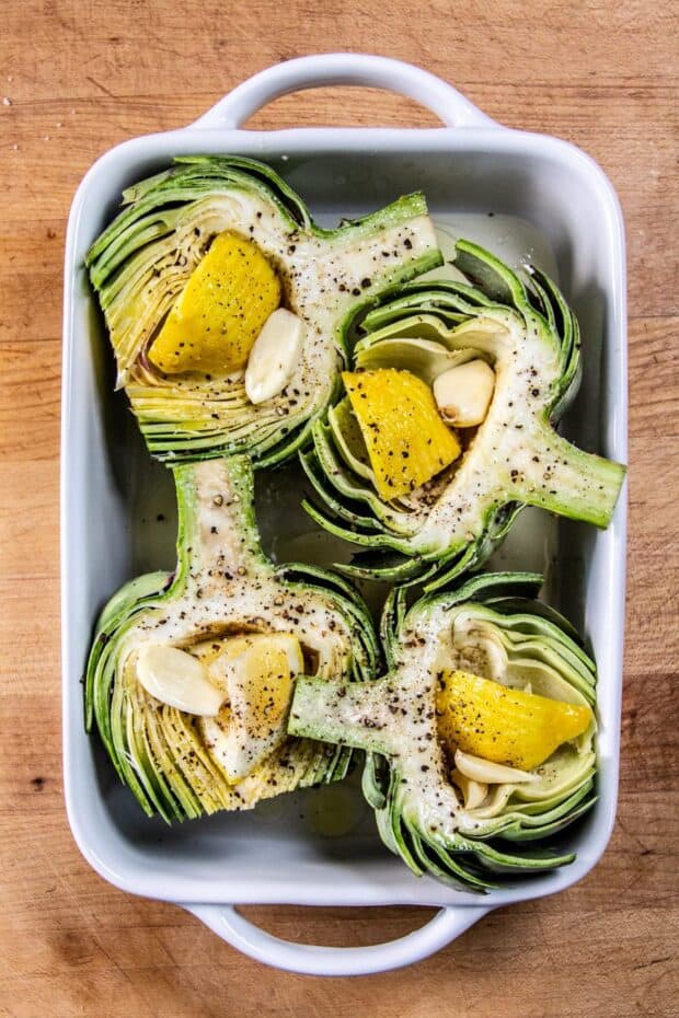 Four raw artichoke halves middle side up stuffed with lemon slices & garlic cloves all placed in a white baking dish.