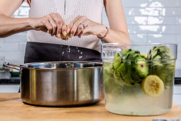 Chef squeezing lemon juice into large silver pot. Artichokes soaking in cold water with lemon halves-set aside