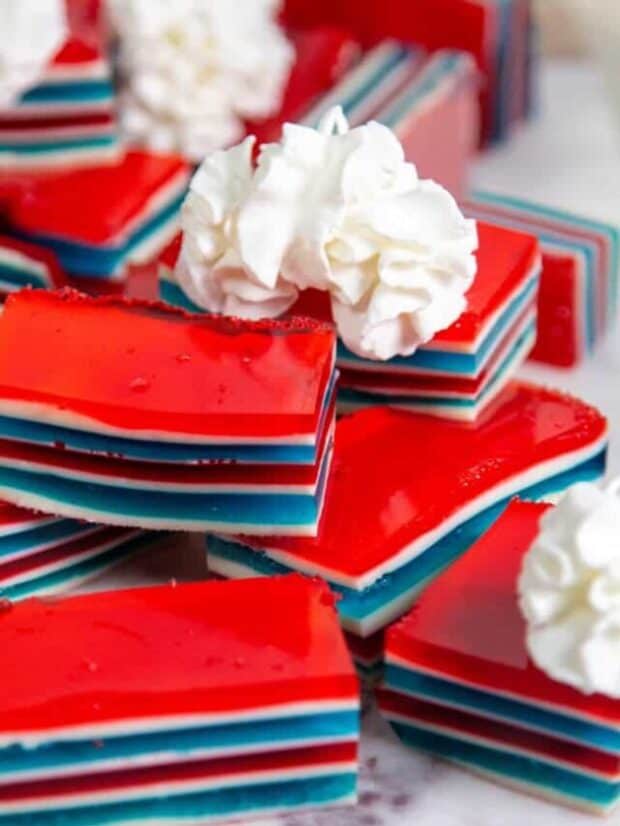 Multiple red, white, and blue stripped jello cake cubes all stacked on a marble slab with a few swirls of whipped cream.